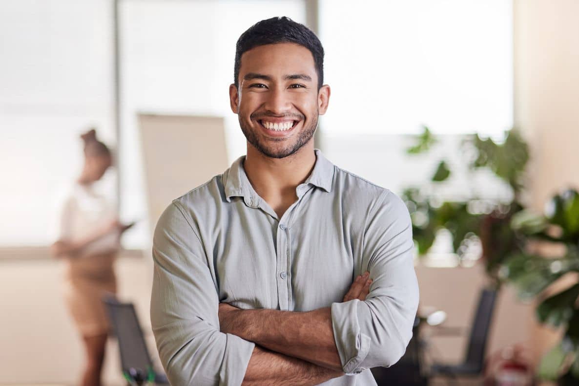 A young man wearing a light gray button up with arms folded smiling